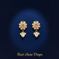 A pair of gold dangle earrings with pink oval stones, surrounded by clear stones and hanging white pearl drops, displayed on a dark blue background with the text 'Rosé Aura Drops' at the bottom.